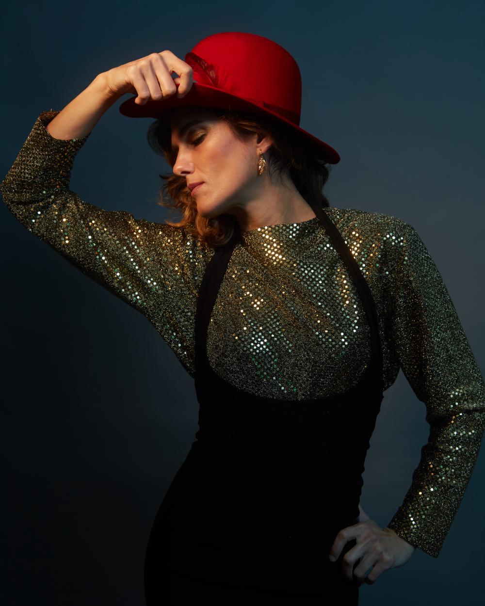 High-fashion editorial portrait of a woman in a shimmering gold sequin top and structured black dress, wearing a statement red felt hat with feather trim, striking a dramatic pose against a cinematic teal backdrop with moody lighting.