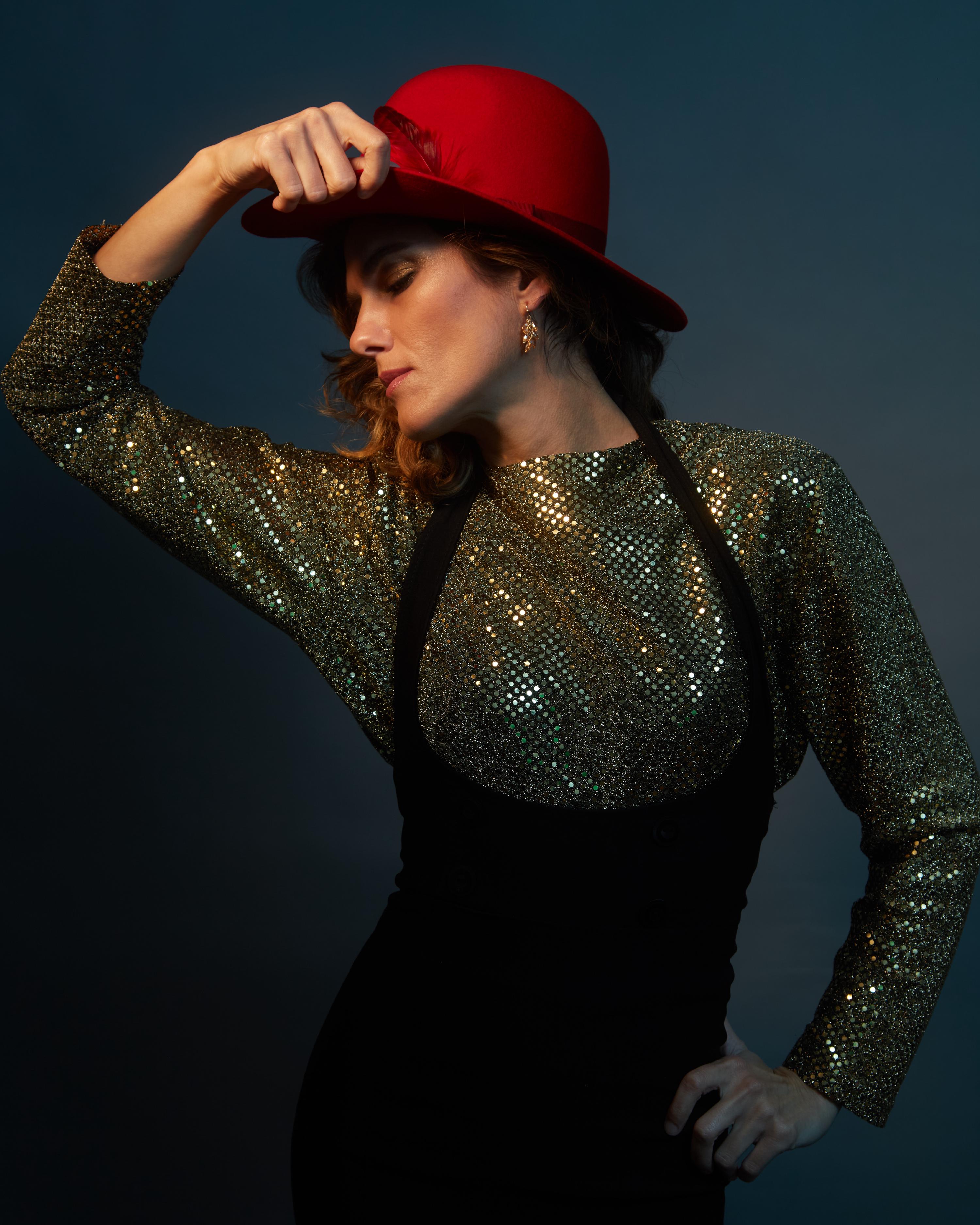High-fashion editorial portrait of a woman in a shimmering gold sequin top and structured black dress, wearing a statement red felt hat with feather trim, striking a dramatic pose against a cinematic teal backdrop with moody lighting.