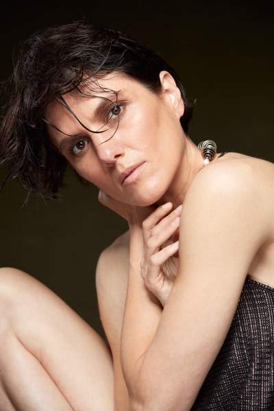Low-key editorial beauty portrait of a woman with short wet dark hair and dewy skin, gazing at the camera with hands at her neck against a dark olive-brown Gravity studio backdrop.