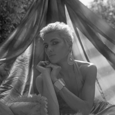 Black and white portrait of a woman with short platinum blonde hair, wearing large statement earrings and a bracelet, posing elegantly with her chin resting on her hands against a flowing fabric backdrop.
