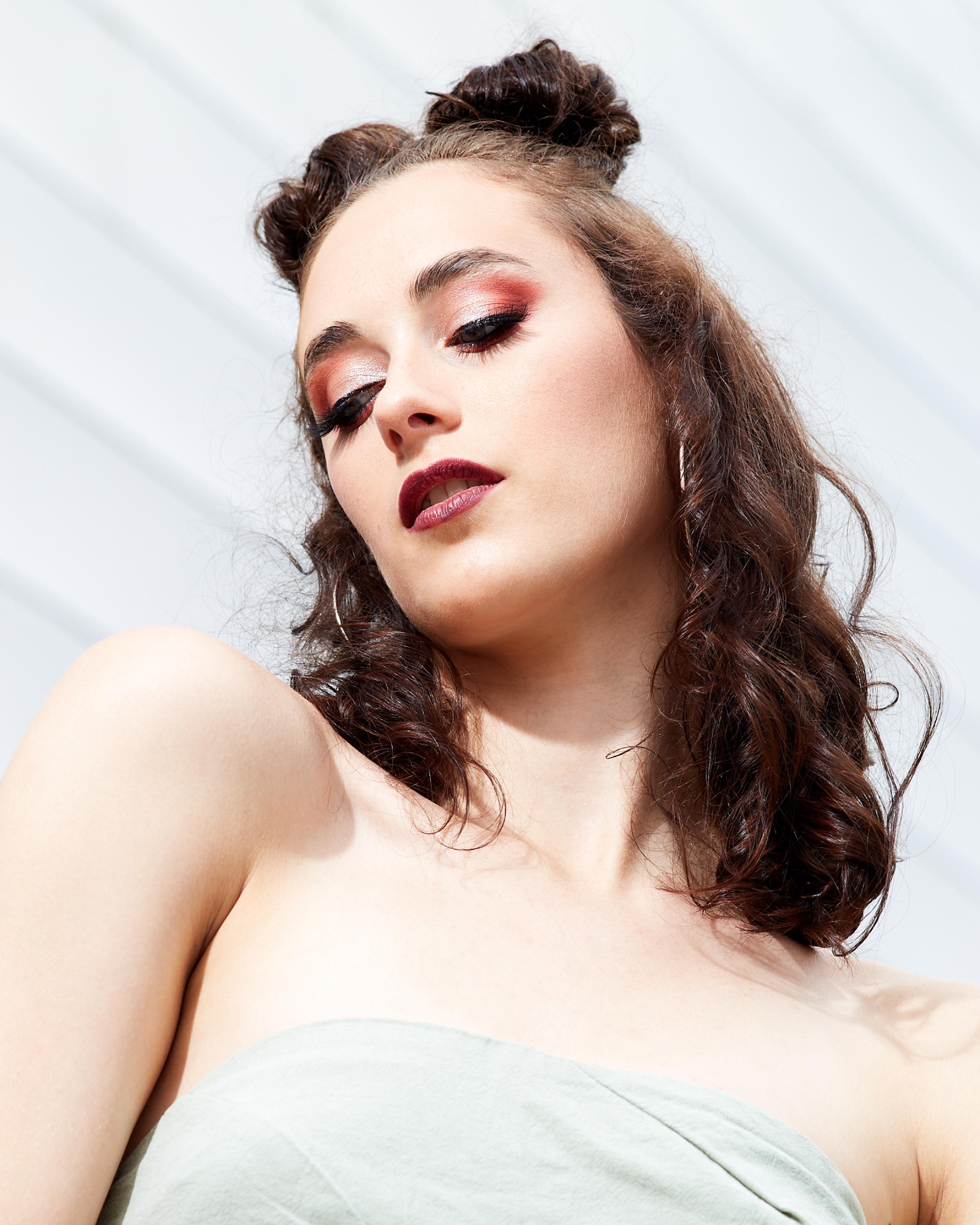 Close-up portrait of a woman with bold makeup, including red eyeshadow and dark lipstick, styled with two high buns and wavy hair, wearing a strapless light green top against a white textured background.