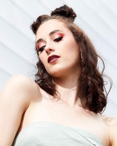 Close-up portrait of a woman with bold makeup, including red eyeshadow and dark lipstick, styled with two high buns and wavy hair, wearing a strapless light green top against a white textured background.